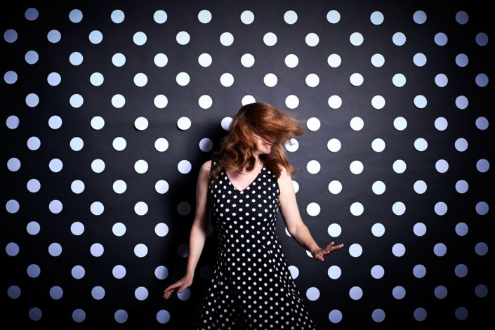 Eine Dame in einem schwarzen Kleid mit weissen Punkten tanzt vor einer schwarzen Wand mit weissen Punkten
