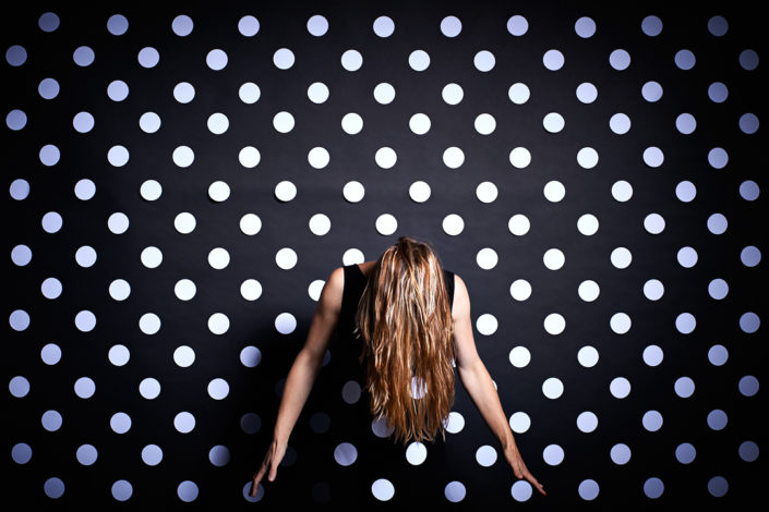 Eine Dame in einem schwarzen Kleid mit weissen Punkten steht vor einer schwarzen Wand mit weissen Punkten
